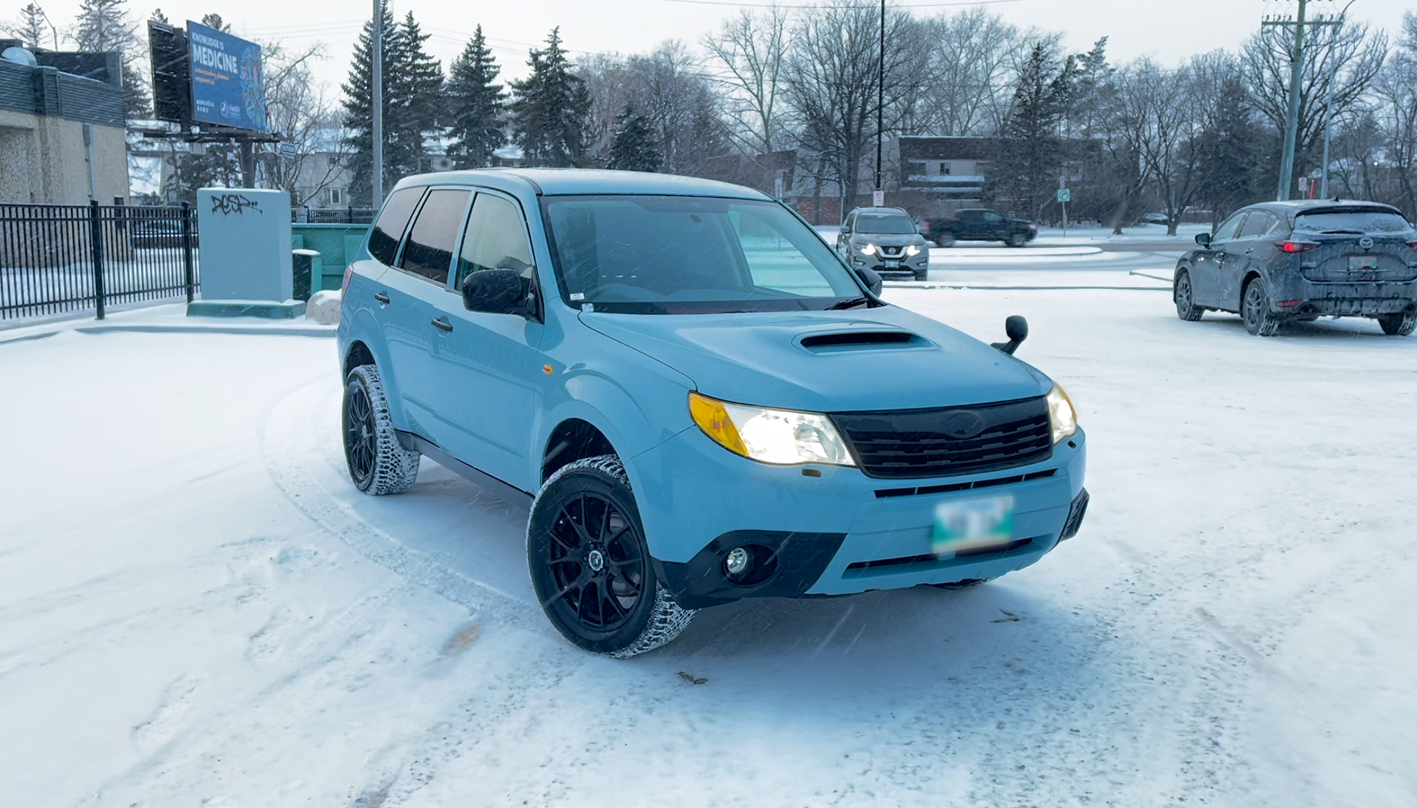 A blue Subaru Forester in winter with snow on the ground in a parking lot.