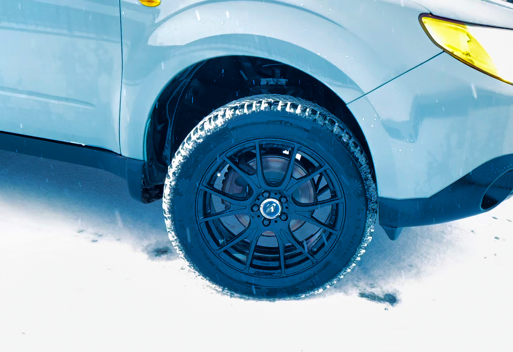 A closeup of a blue Subaru Forester tire in winter with snow on the ground in a parking lot.