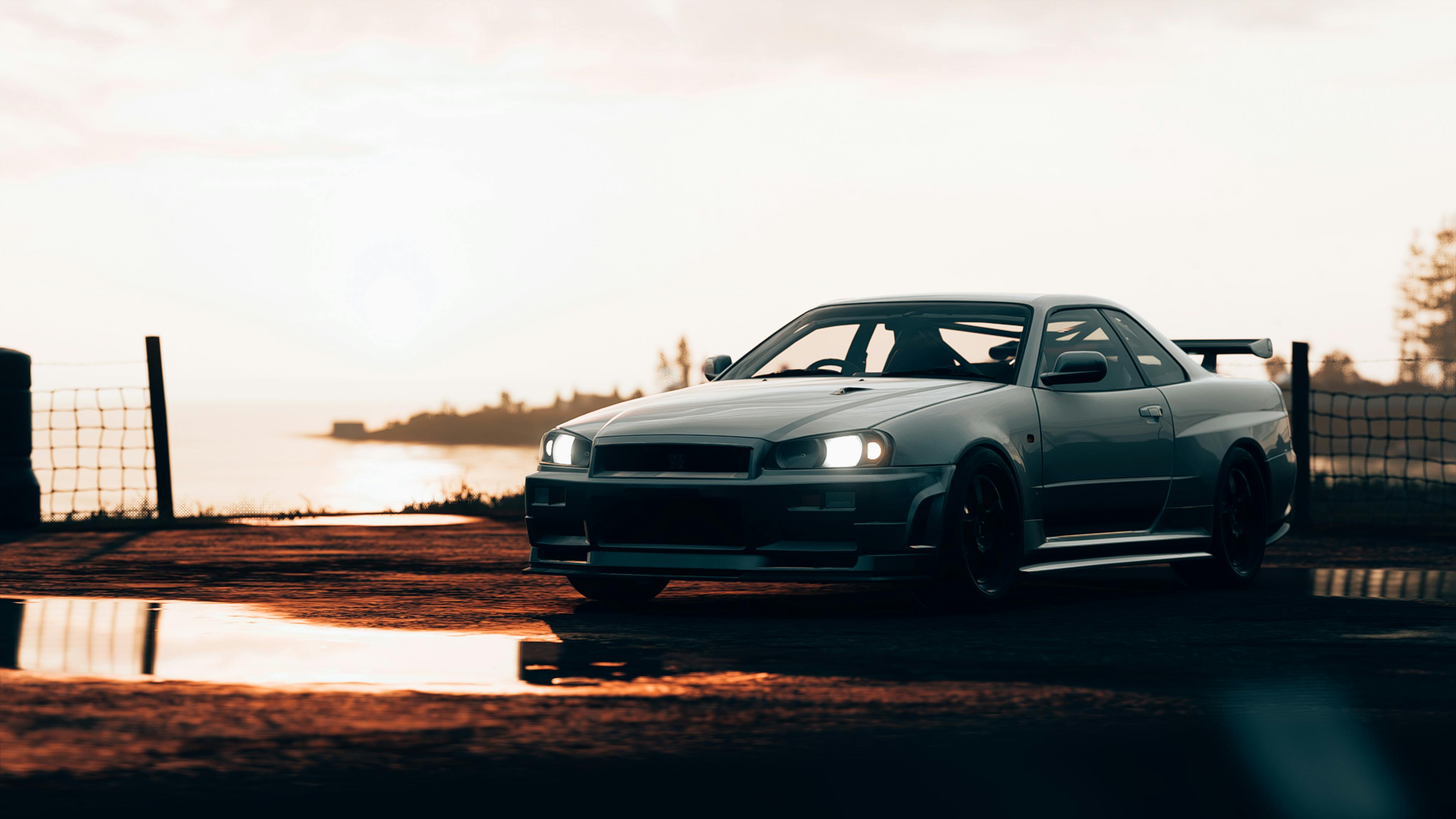 A grey Nissan Skyline in front of a lake with it's headlights on at dusk.