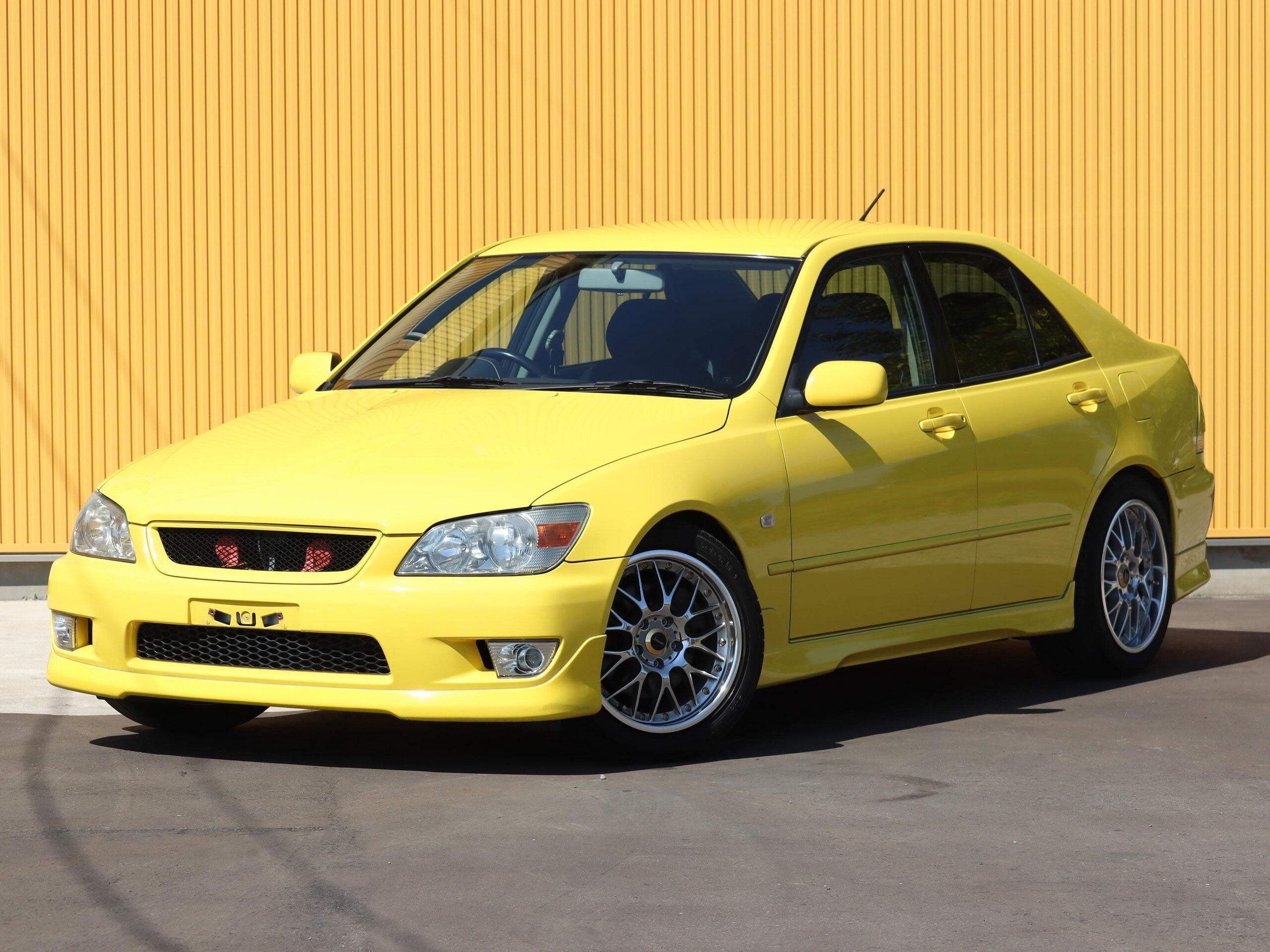 A yellow Toyota Altezza RS200 in front of a yellow wall. This is a JDM car that can be imported into the USA and Quebec in 2025.