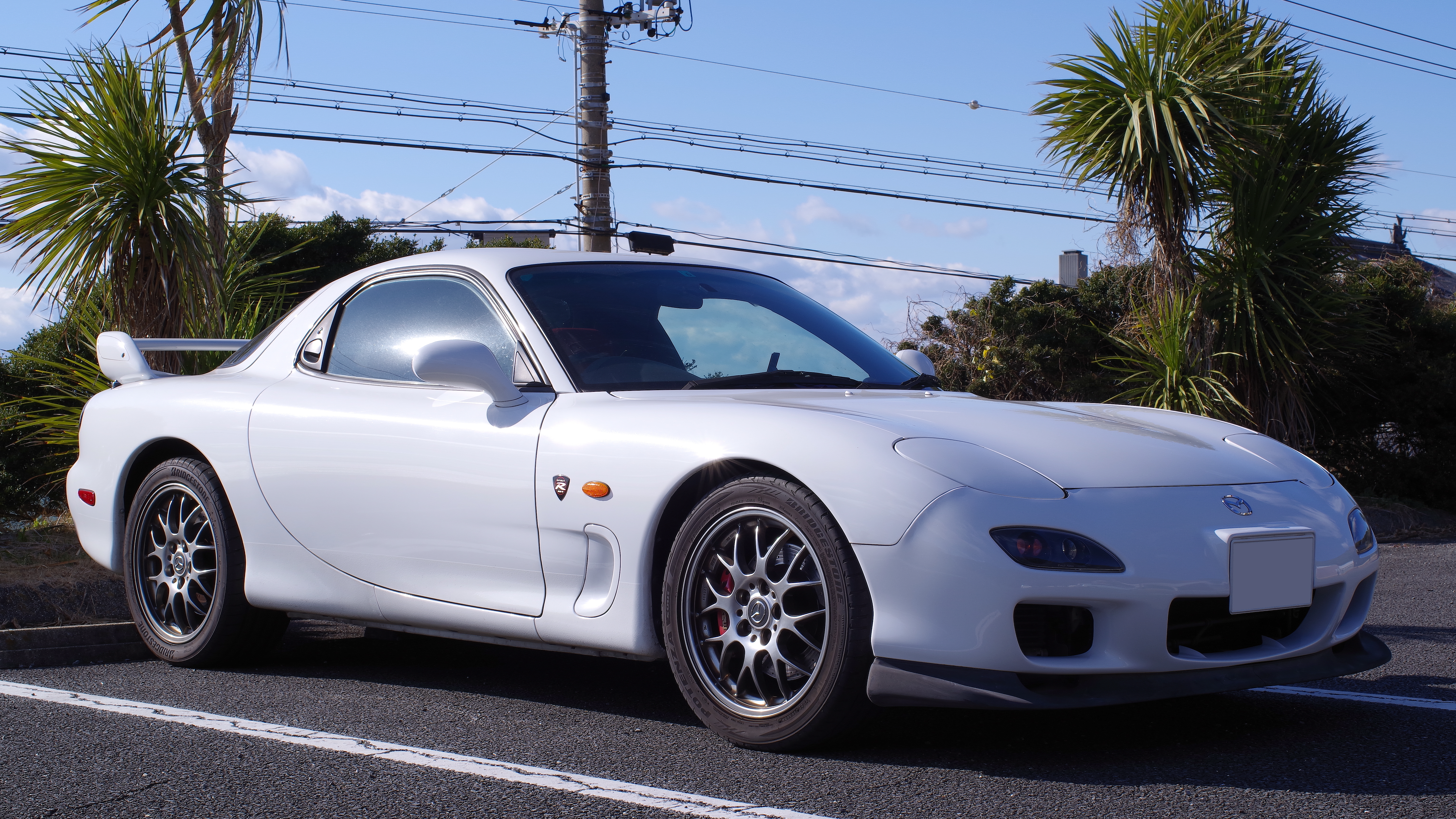A white Mazda RX-7 Spirit R (FD3S) in front of some palm trees and a telephone line. This is a JDM car that can be imported into the USA and Quebec in 2025.