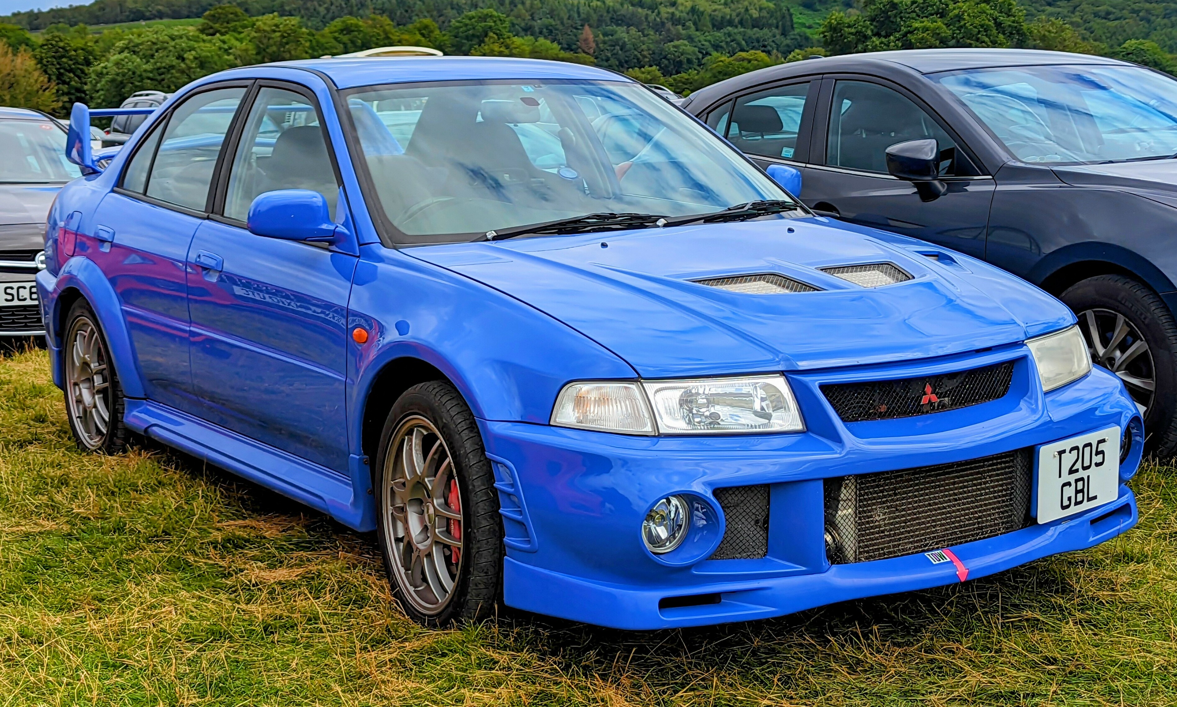A bright blue Mitsubishi Lancer evolution parked on some grass amongst other vehicles. This is a JDM car that can be imported into the USA and Quebec in 2025.