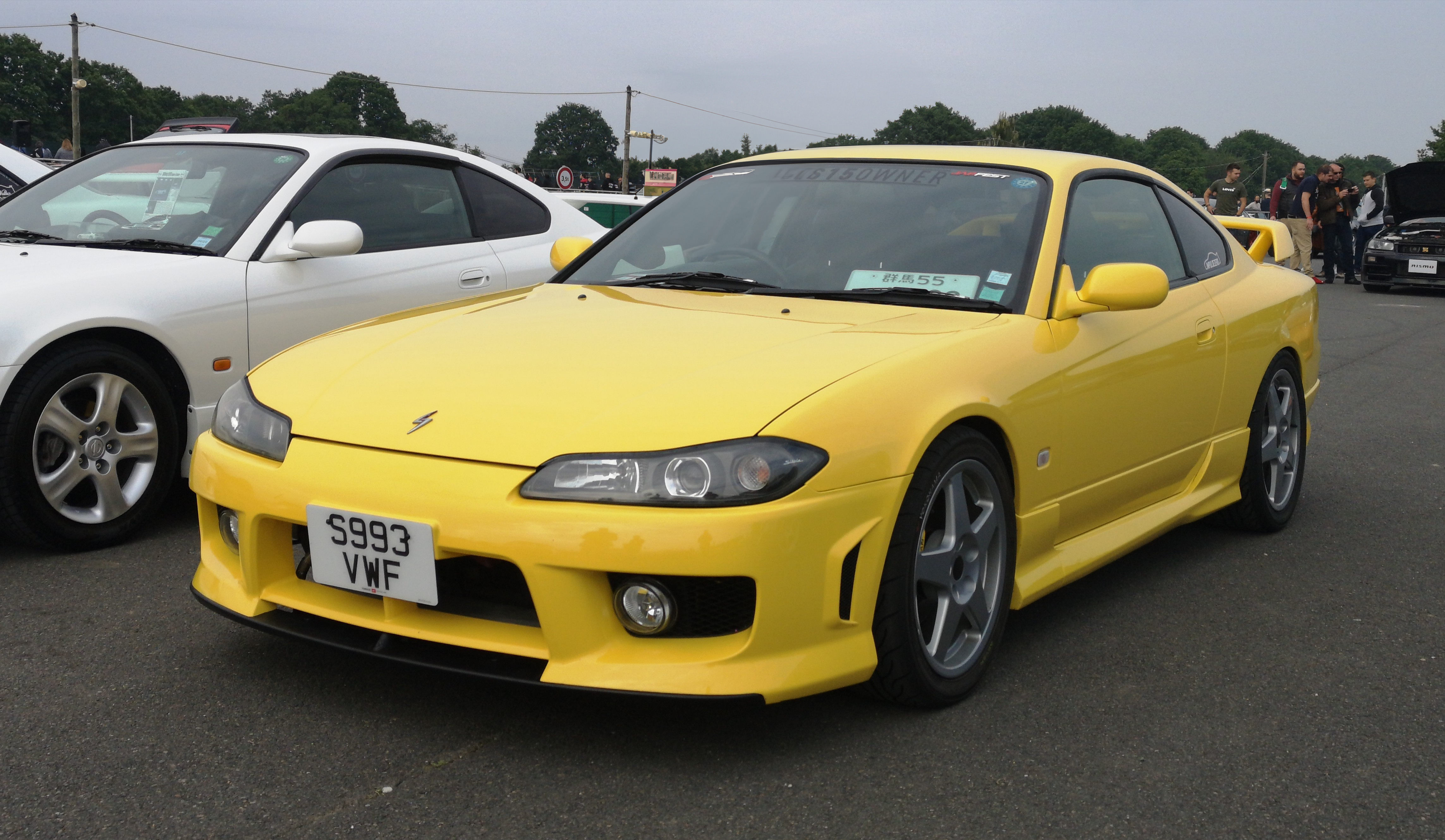 A yellow Nissan Silvia S15 Spec-R parked next to a white vehicle with trees in the background. This is a JDM car that can be imported into the USA and Quebec in 2025.