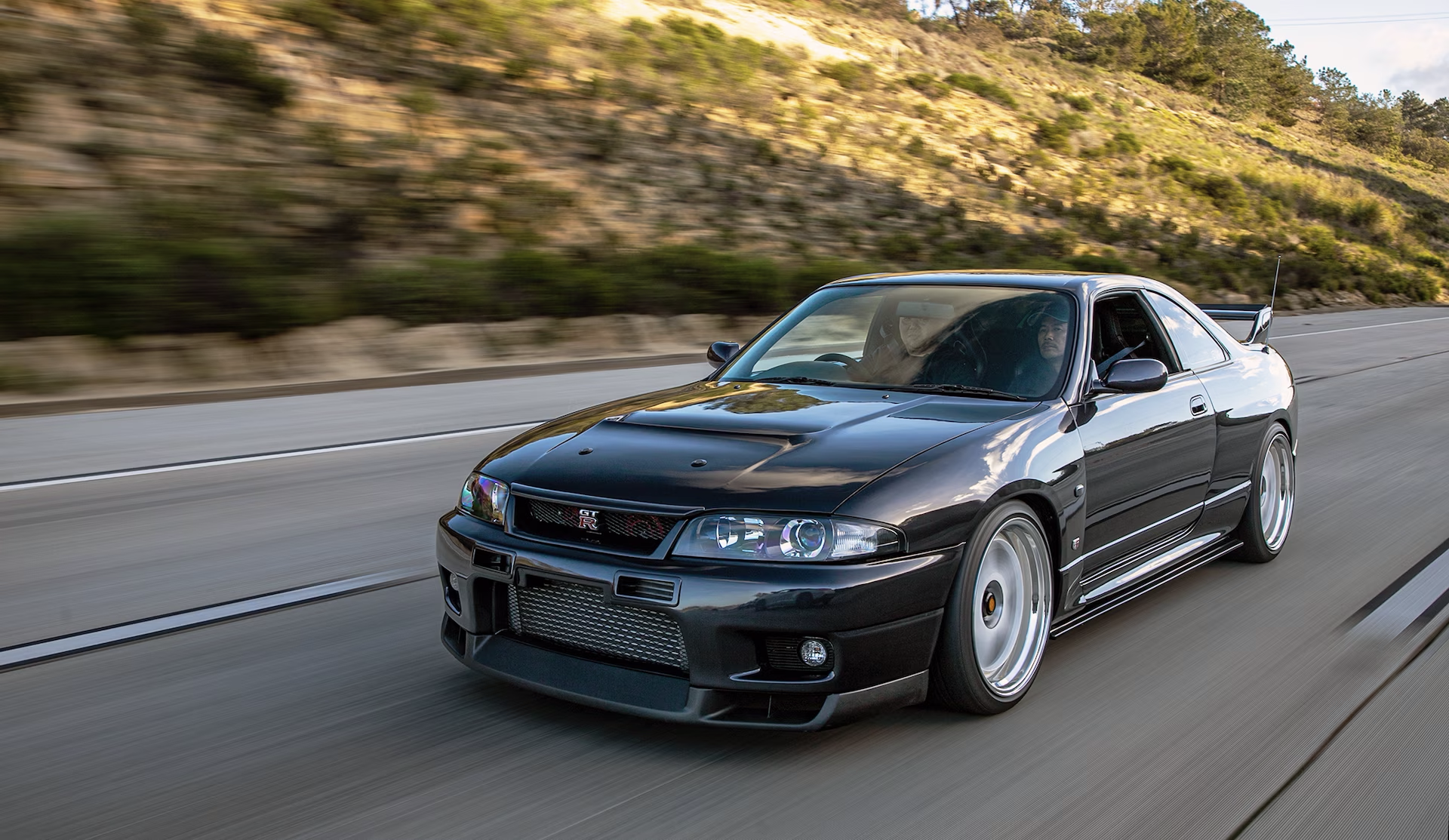 A black Nissan Skyline GTR in motion on a highway running past a hillside. This is a JDM car that can be imported in the USA and Quebec in 2024.