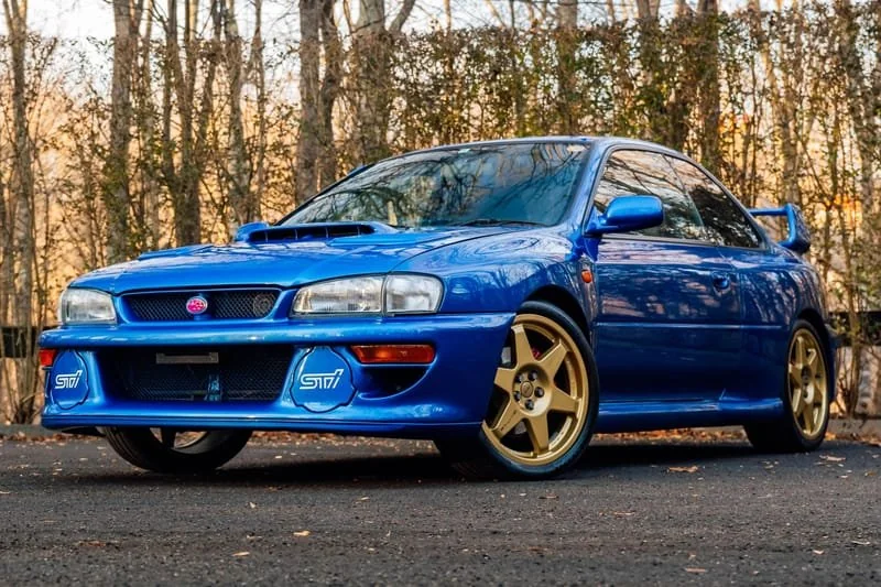 A bright blue Subaru Impreza WRX STI parked in front of a line of trees. This is a JDM car that can be imported in the USA and Quebec in 2024.