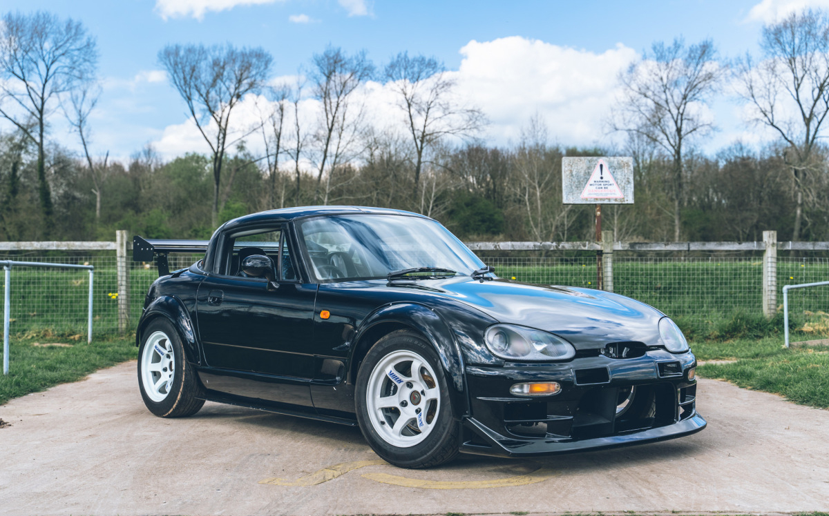 A black Suzuki Cappucino parked in front of a wire fence with a field of grass and treeline behind it. This is a JDM car that can be imported in the USA and Quebec in 2024.