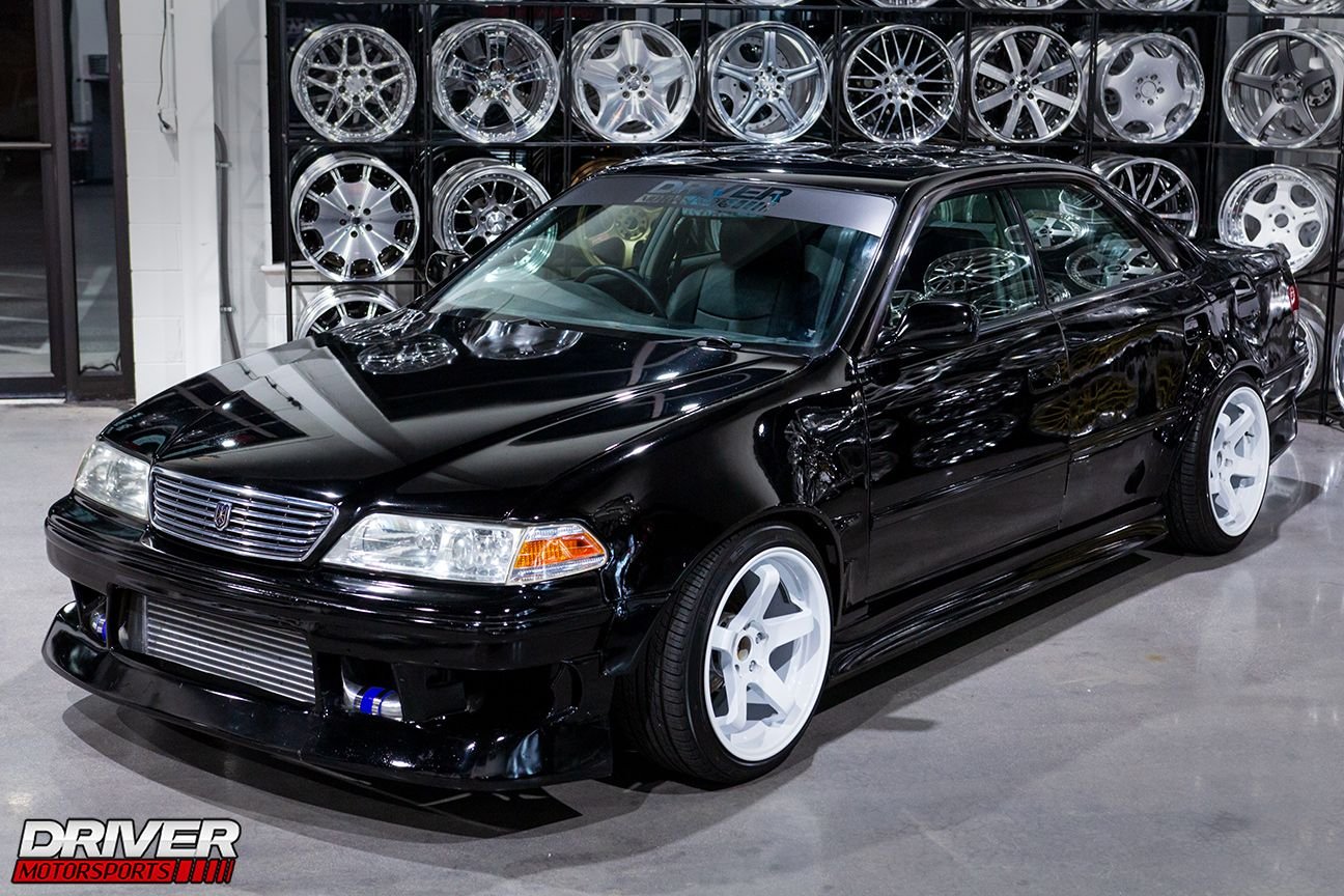 A black Toyota Mark II (JZX100) in a showroom in front of a wall of car rims. This is a JDM car that can be imported in the USA and Quebec in 2024.