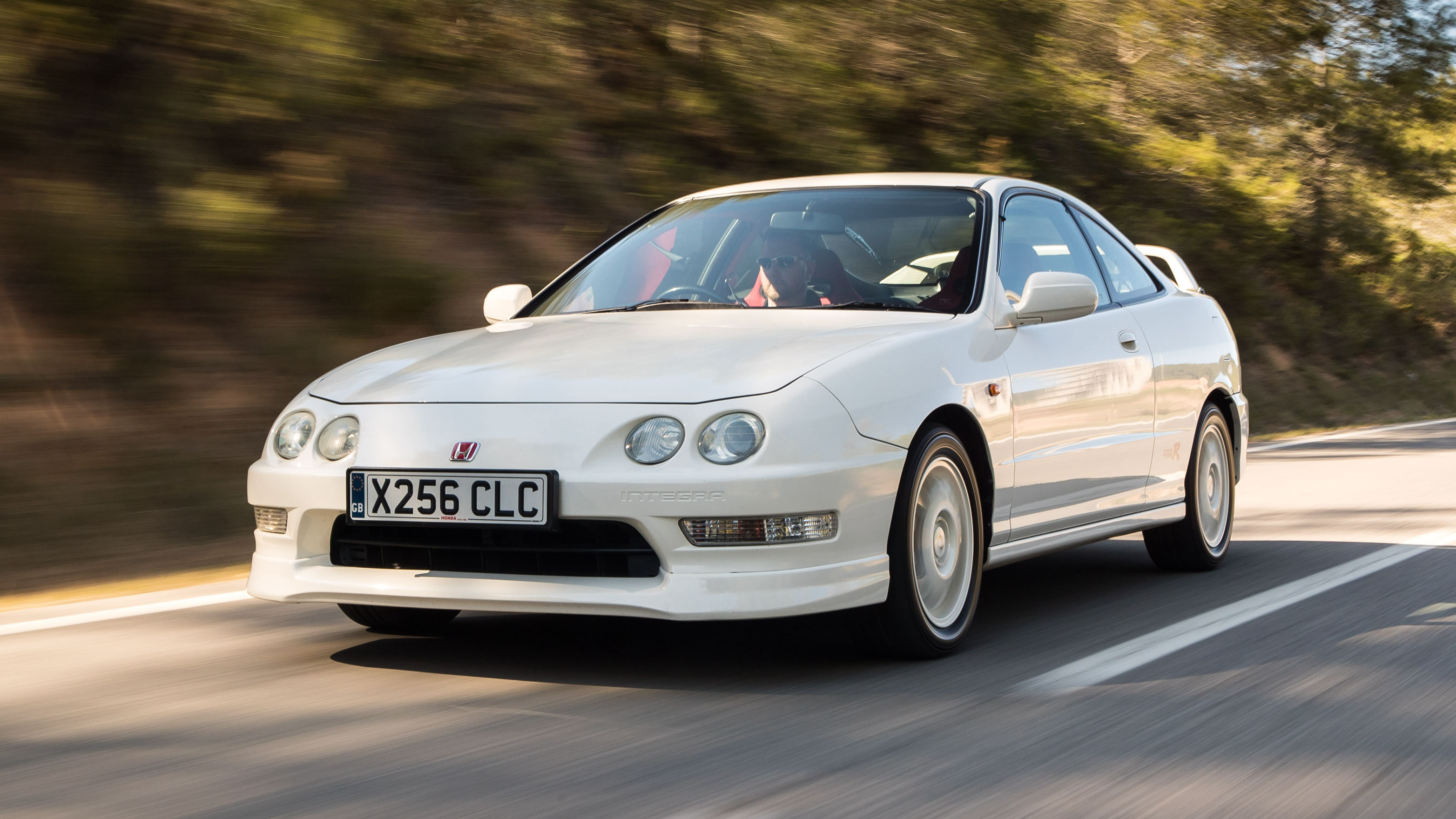 A white Honda Integra Type R speeding down a highway in front of a line of trees. This is a JDM car that can be imported in the USA and Quebec in 2024.